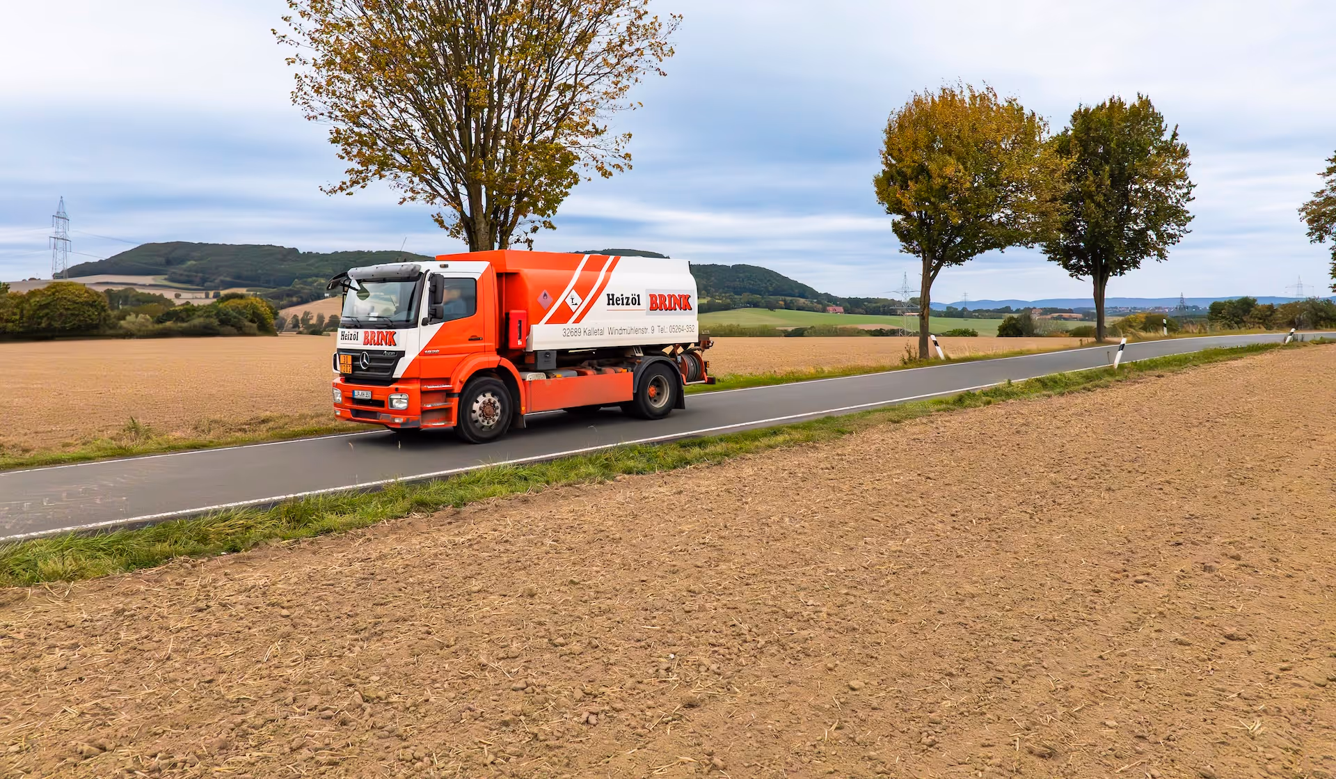Ein rotes Heizöllieferfahrzeug fährt auf einer Landstraße mit Feldern und Bäumen im Hintergrund.